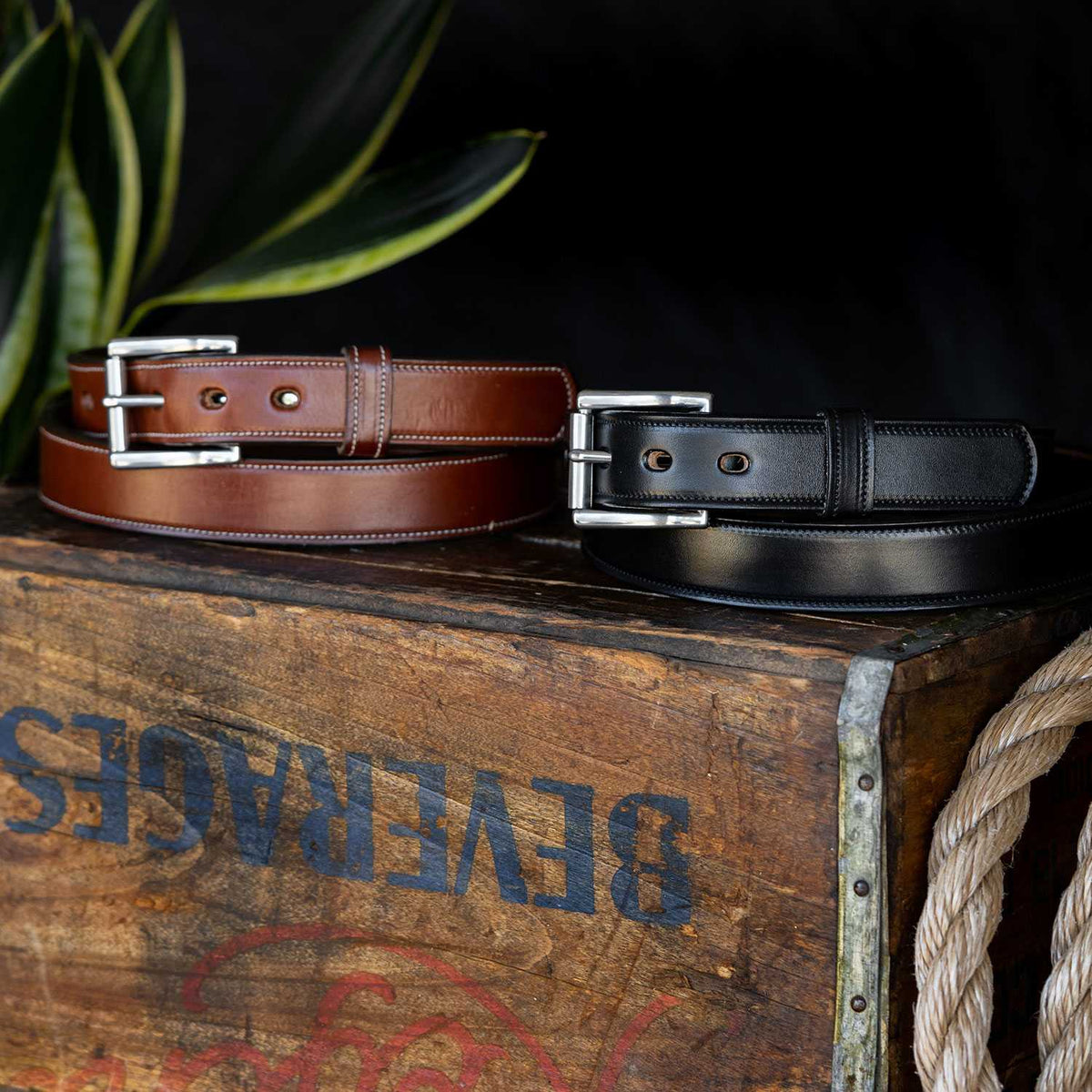 Two "The Highland Lined" belts by Hanks Belts—one brown, one black—crafted from English bridle leather with silver buckles, rest on a rustic wooden box accented with a rope and green leaves.