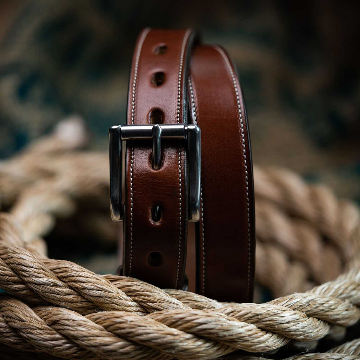 The Highland Lined belt by Hanks Belts, made from English bridle leather with a shiny silver buckle, is shown coiled atop thick rope against a blurred background.