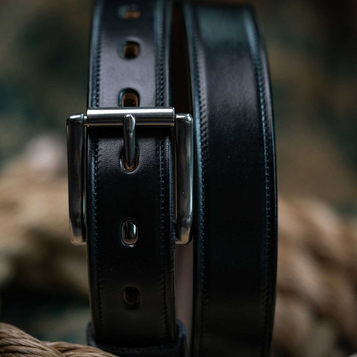 Close-up of The Highland Lined by Hanks Belts, crafted from English bridle leather with a silver buckle, detailed stitching, and multiple adjustment holes. The blurred background displays earthy tones.