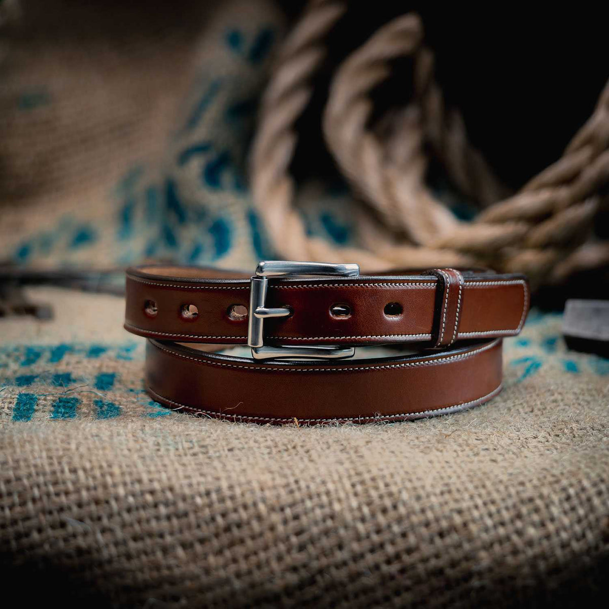The Highland Lined by Hanks Belts, made from vegetable-tanned leather and finished with a silver buckle, is coiled on textured burlap, with thick rope and blue markings creating a rustic background.