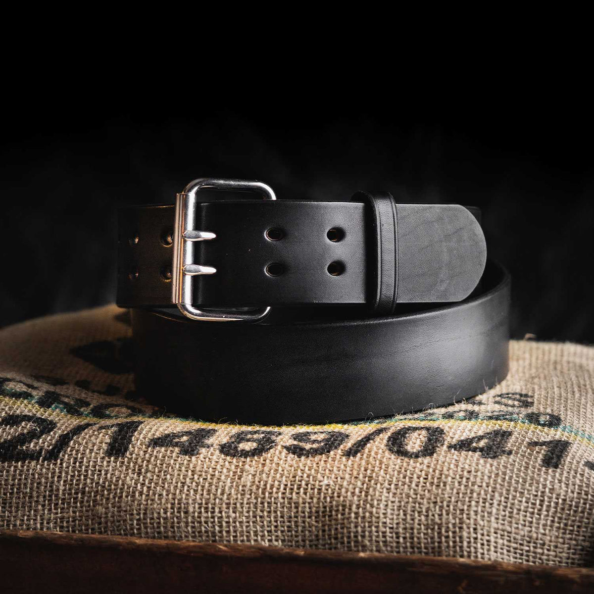 The Marshall by Hanks Belts, a black leather Garrison Style Belt with a silver buckle, is coiled atop a burlap sack with printed text against a dark background.