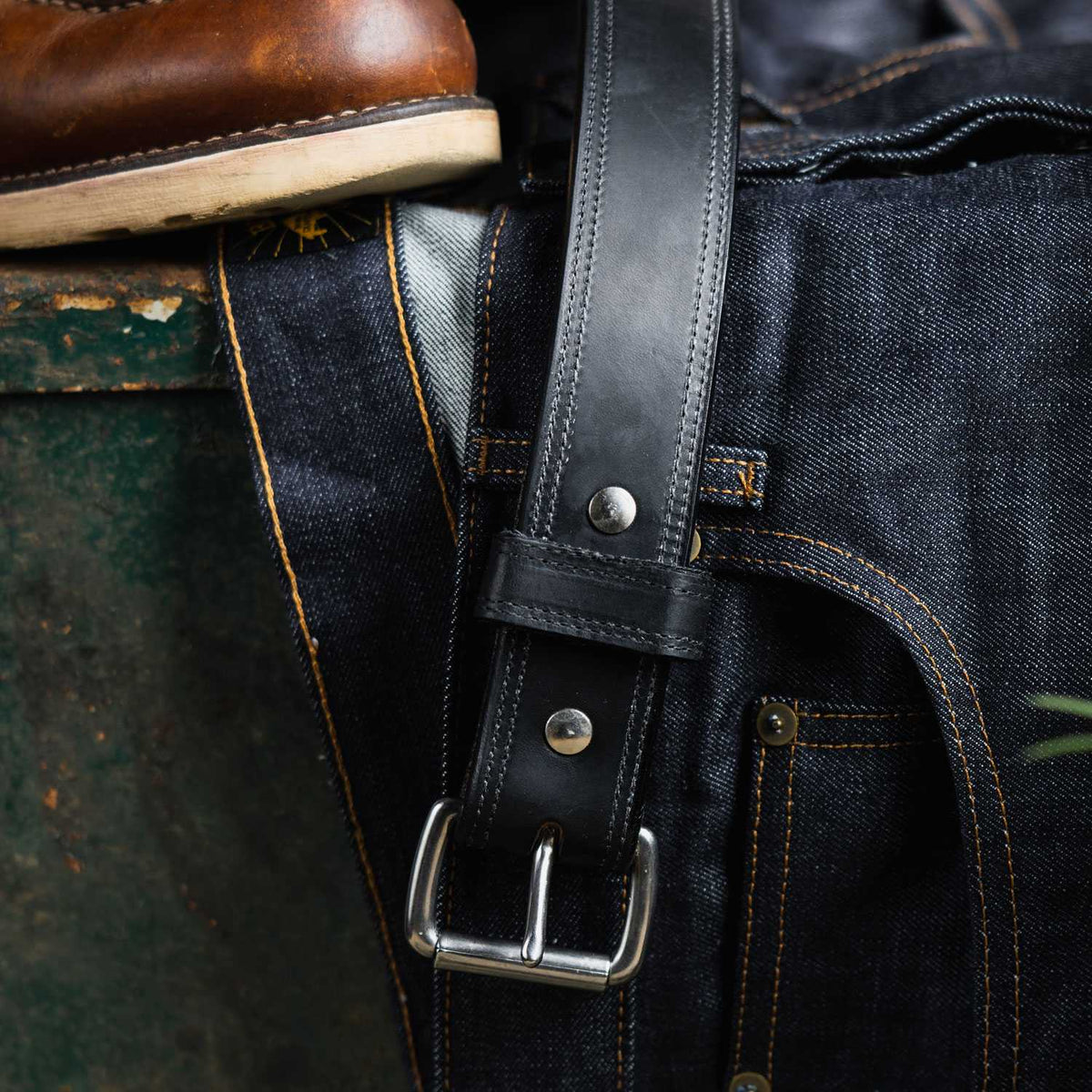 A close-up of The Old World Harness Leather Belt - 1.5" by Hanks Belts, made from black full-grain USA-tanned Hermann Oak leather with a silver buckle, resting on dark blue denim jeans beside a brown leather shoe.