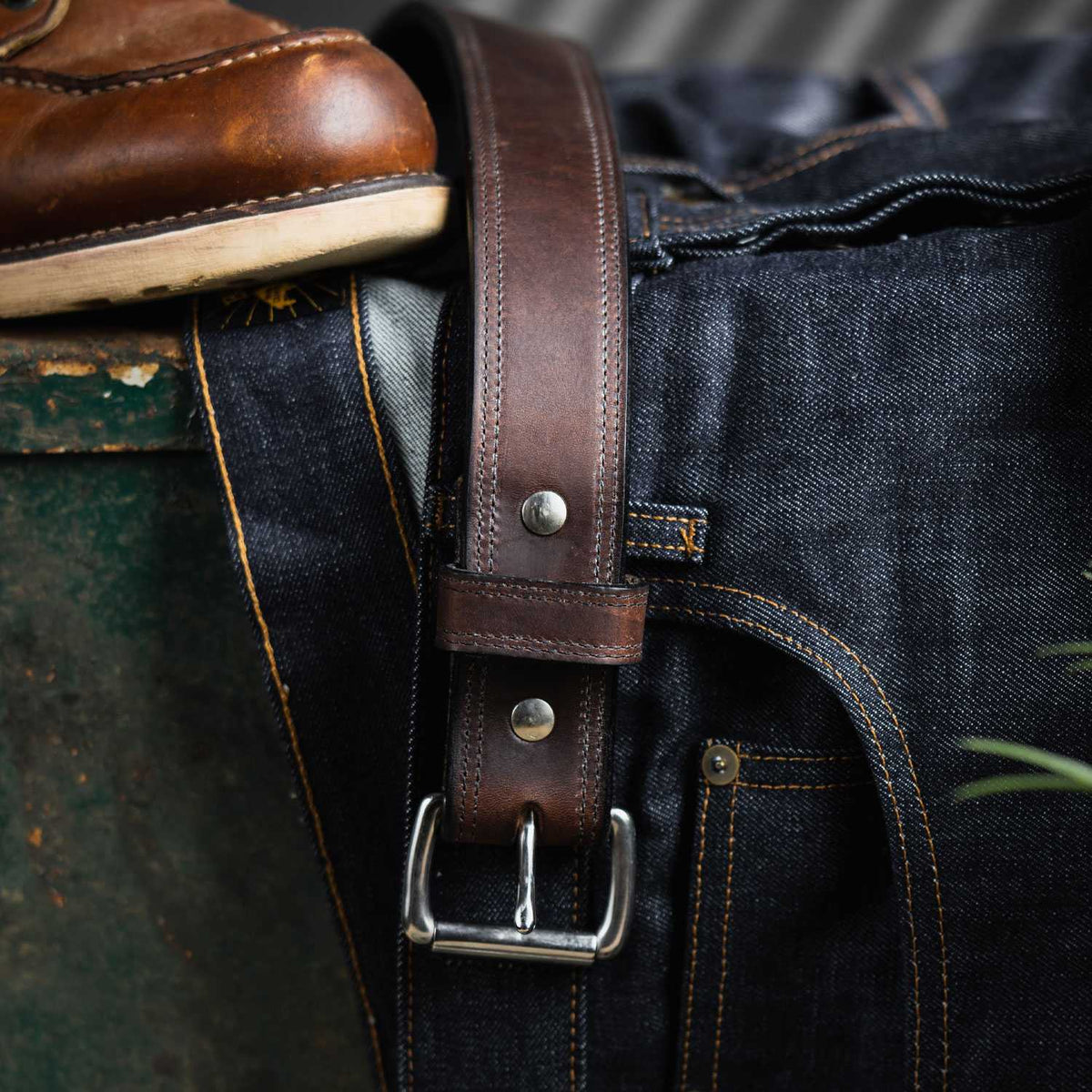 A Hanks Belts Old World Harness Leather Belt - 1.5" is threaded through the loops of dark blue denim jeans, with a brown leather shoe nearby on a green surface.