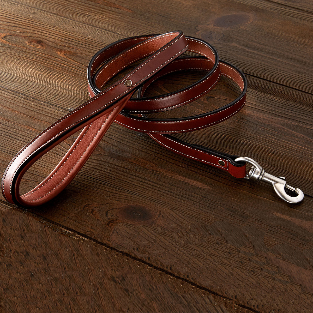 The Rover Standard Leash by Hanks Belts, made from brown English Bridle leather with white stitching, a metal clasp, and padded handle, lies coiled on a wooden surface.