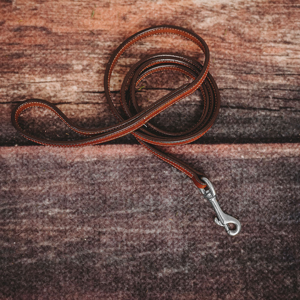 The Hanks Belts Rover Standard Leash, crafted from coiled brown English Bridle leather and featuring a metal clasp, rests on a rustic wooden surface.
