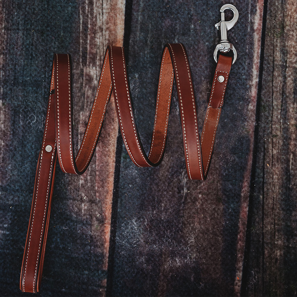 The Hanks Belts Rover Standard Leash, made from brown English Bridle leather with white stitching, a padded handle, and silver clasp, is arranged in a zigzag on a dark, textured wooden surface.