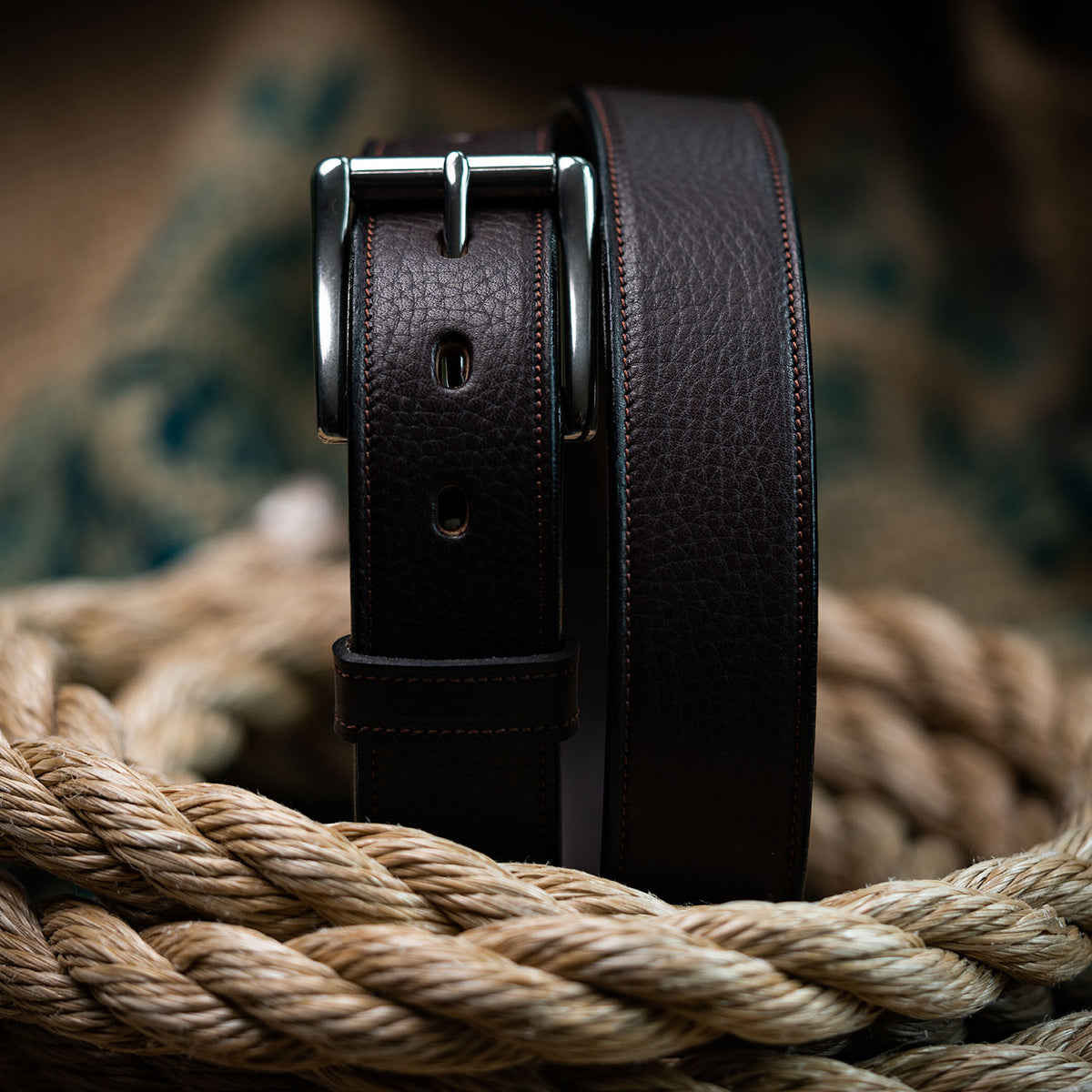 The Tuscan Embossed Lined by Hanks Belts, crafted from dark brown English Bridle Leather with a stainless steel buckle, is coiled on thick ropes against a blurred fabric backdrop.