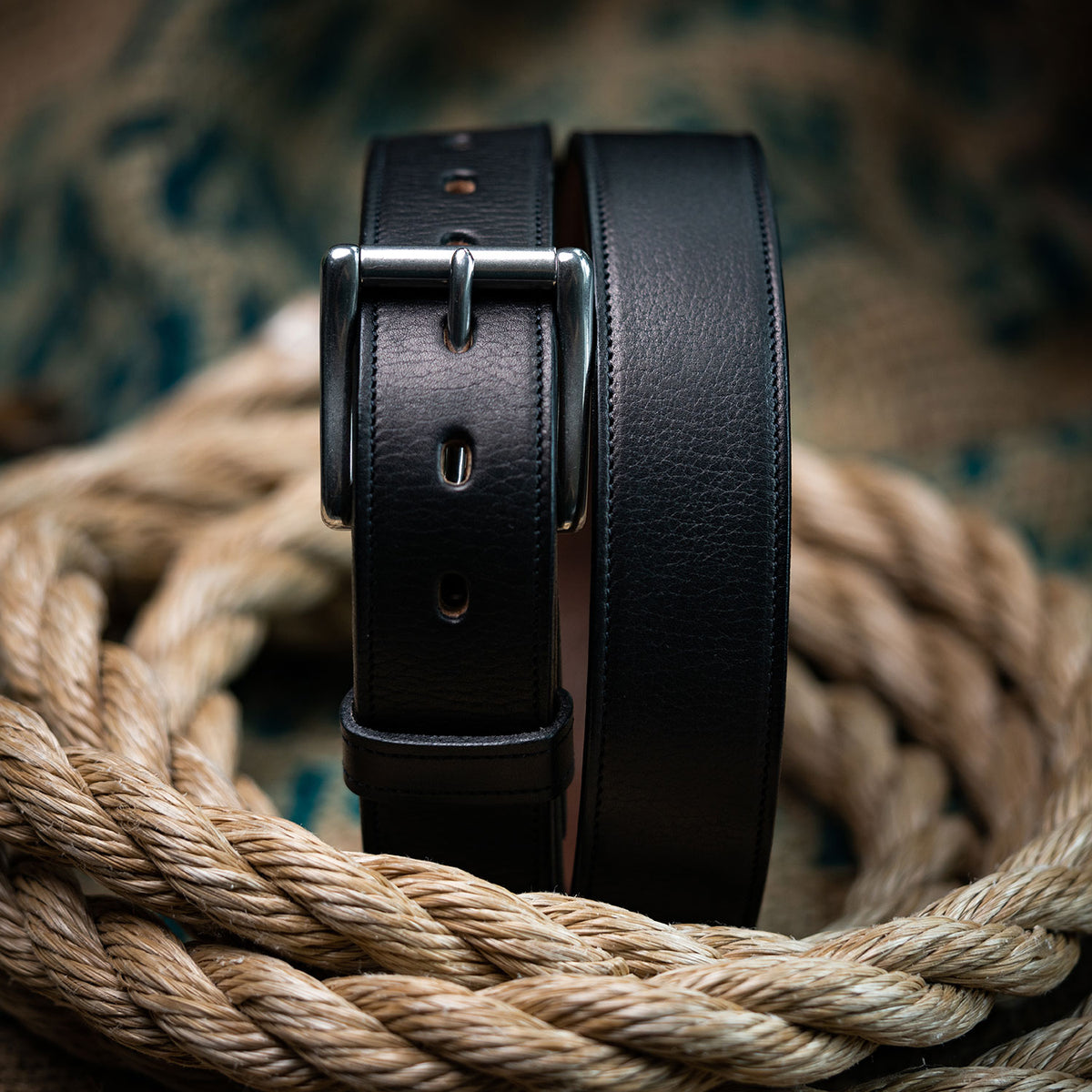 The Tuscan Embossed Lined by Hanks Belts is coiled atop thick tan rope, its premium leather and stainless steel buckle highlighted against a blurred decorative background.