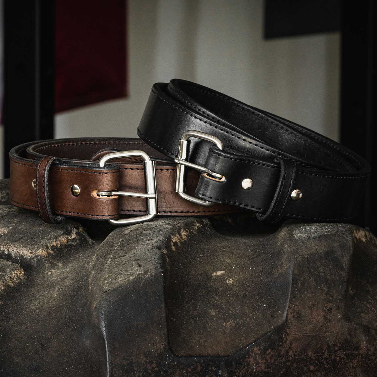 Close-up of two Hanks Belts Xtreme Belts - 1.75" draped over a textured stone surface. One belt is brown with a silver buckle, and the other is black with a silver buckle. Both heavy-duty leather belts feature double prong buckles and stitching details along the edges.
