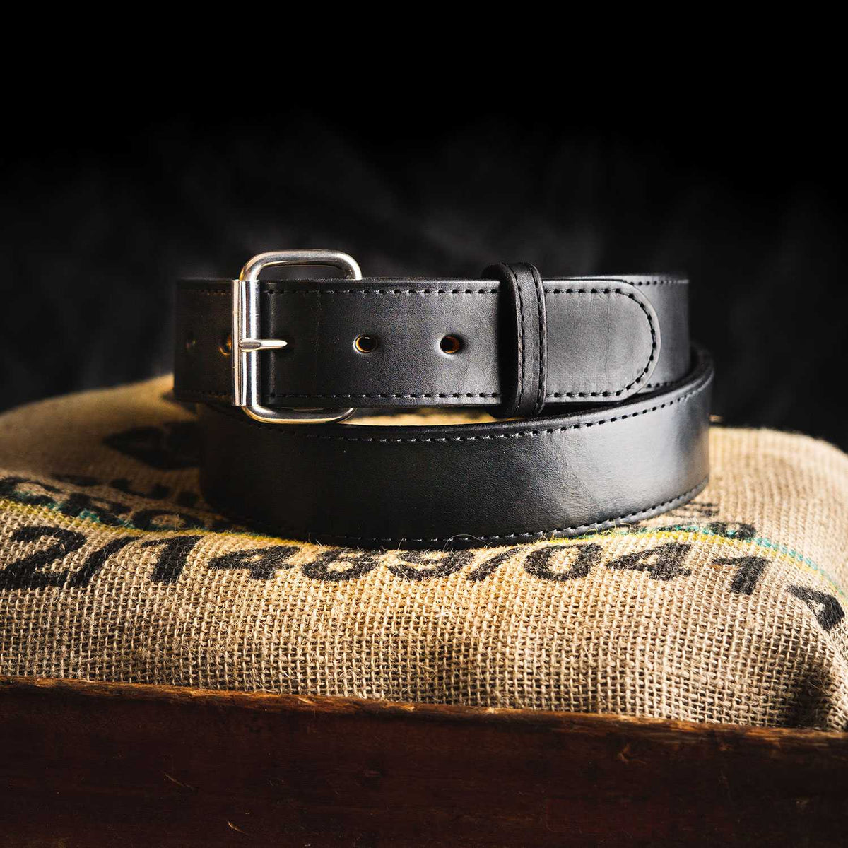 The Xtreme Belt - 1.75" from Hanks Belts, a black heavy-duty leather belt with a silver buckle, is coiled neatly on a piece of burlap fabric that rests atop a wooden surface. The dark background emphasizes the work belt as the main focus of the image.