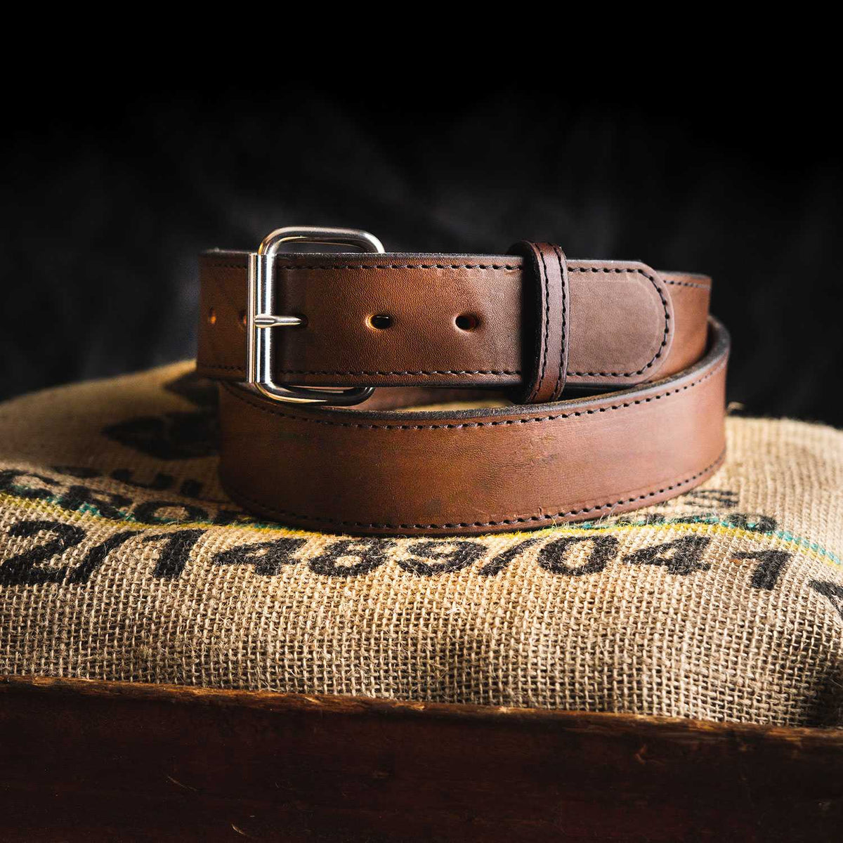 A rolled-up Xtreme Belt - 1.75" from Hanks Belts, featuring a sturdy leather build and a silver buckle, is displayed on burlap fabric. The dark background accentuates the texture and craftsmanship of this durable work belt.