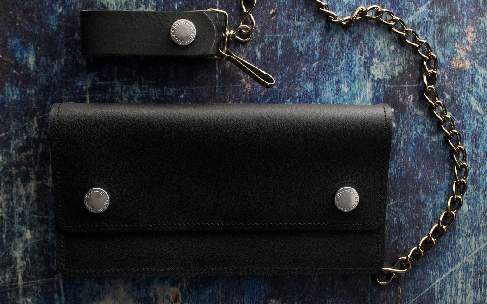 A black leather wallet with silver buttons and a matching keychain, both attached to a gold chain, displayed on a textured blue surface.