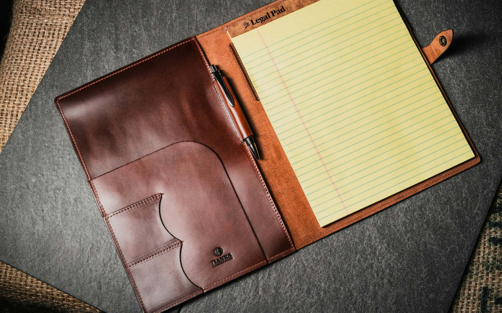 A brown leather portfolio with pockets and a pen holder, open to reveal a yellow lined legal pad on the right, resting on a dark surface with woven fabric visible underneath.