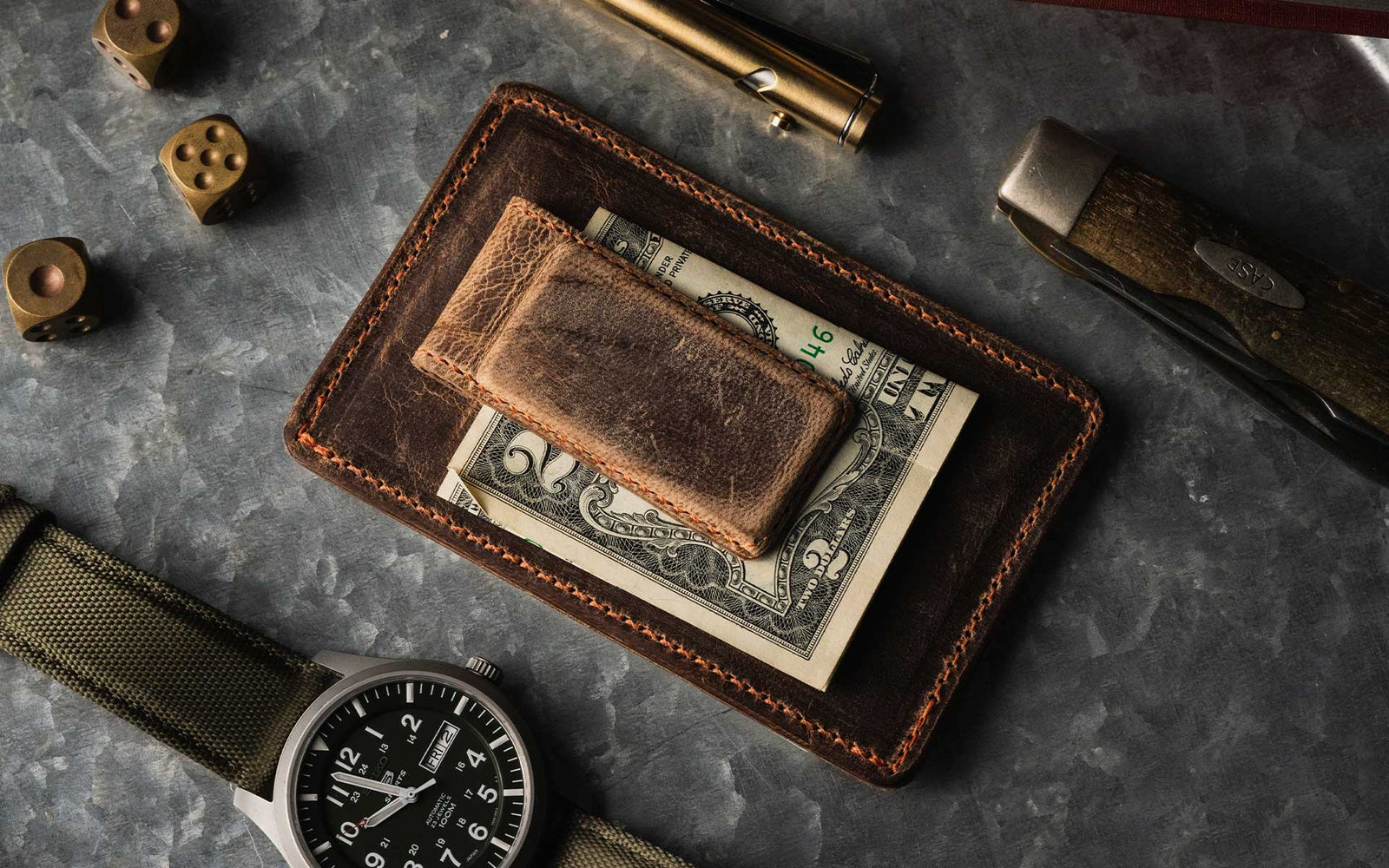 A leather wallet with a money clip holding dollar bills, surrounded by a wristwatch, a knife, brass dice, and a pen on a gray textured surface.