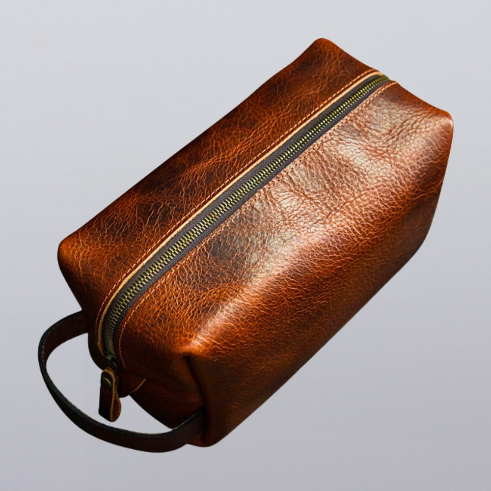 The Hanks Belts Bison Leather Dopp Bag features a durable brown grain texture, secure brass zipper, and a handy carrying loop, all crafted with care and set against a plain light gray background.