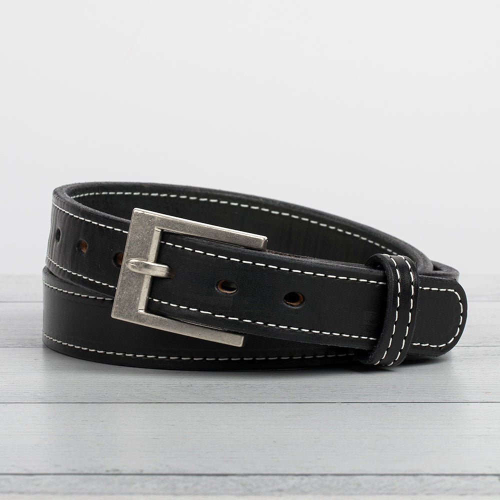 The Bonnie by Hanks Belts is a black full-grain vegetable tanned leather belt with white stitching and a silver rectangular buckle, shown coiled on a light gray wooden surface against a plain white background.