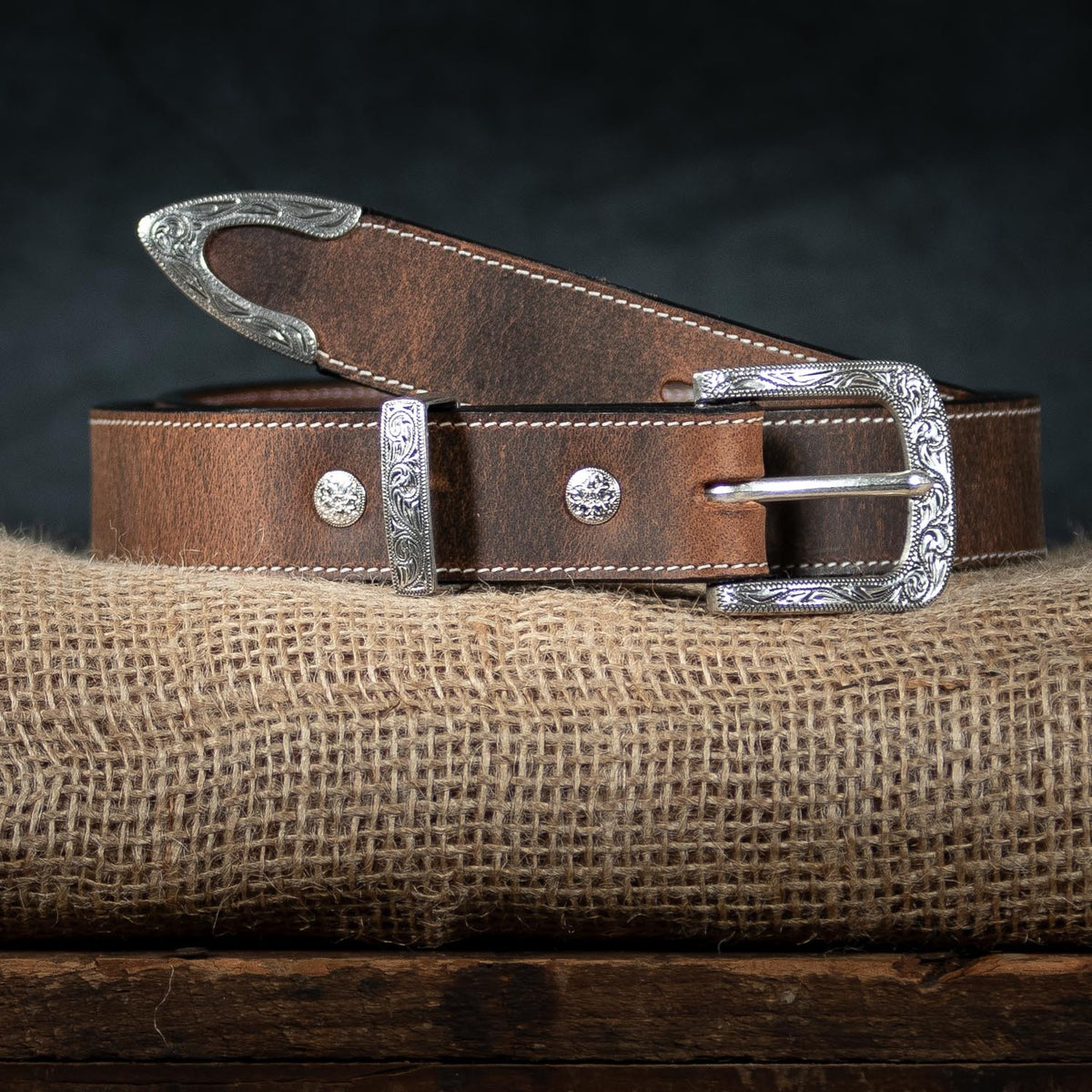 The Dakota Bison by Hanks Belts: a brown North American bison leather western belt with a floral buckle, engraved silver tip, and accents, shown coiled on burlap against a dark background.