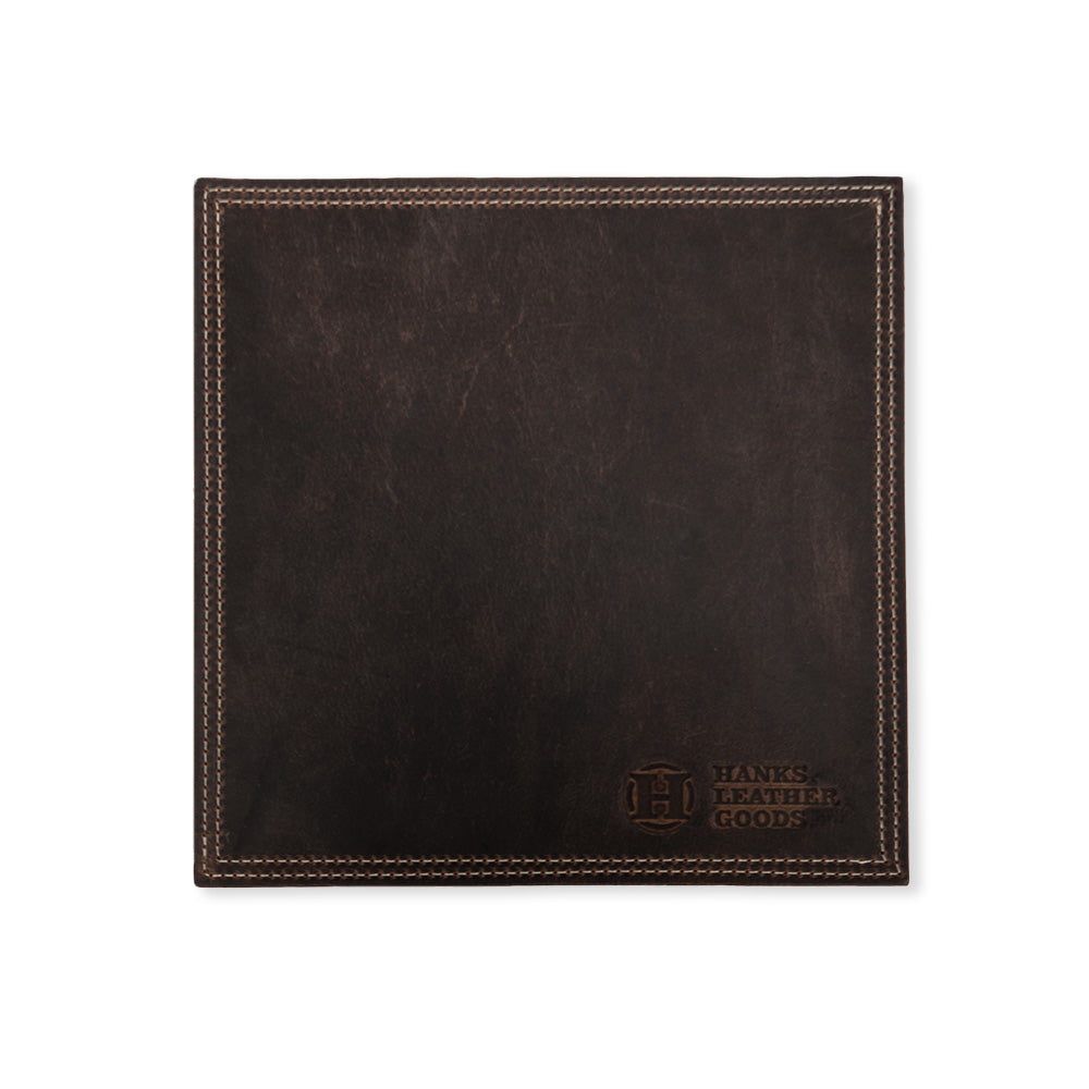A square piece of luxurious dark brown bison leather showcasing visible stitching along its edges, this mouse pad is elegantly embossed in the bottom right corner with "Hanks Belts" and a circular logo featuring the letter "H". Handmade in the USA, it's a refined accessory that enhances any workspace.