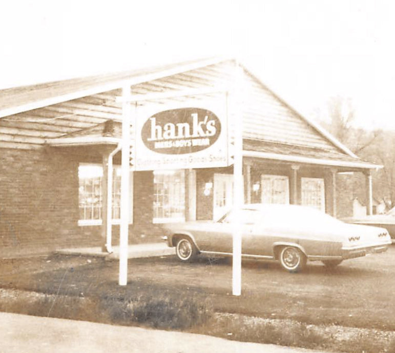Mid 60's .Vintage photo of a building with 'hank's' sign and a car parked in front. Text below the image: With the growth of the business and working 12-14 hour days, sights were set on expansion into a new facility. A new building and addition a year later gave room to expand his offerings. 