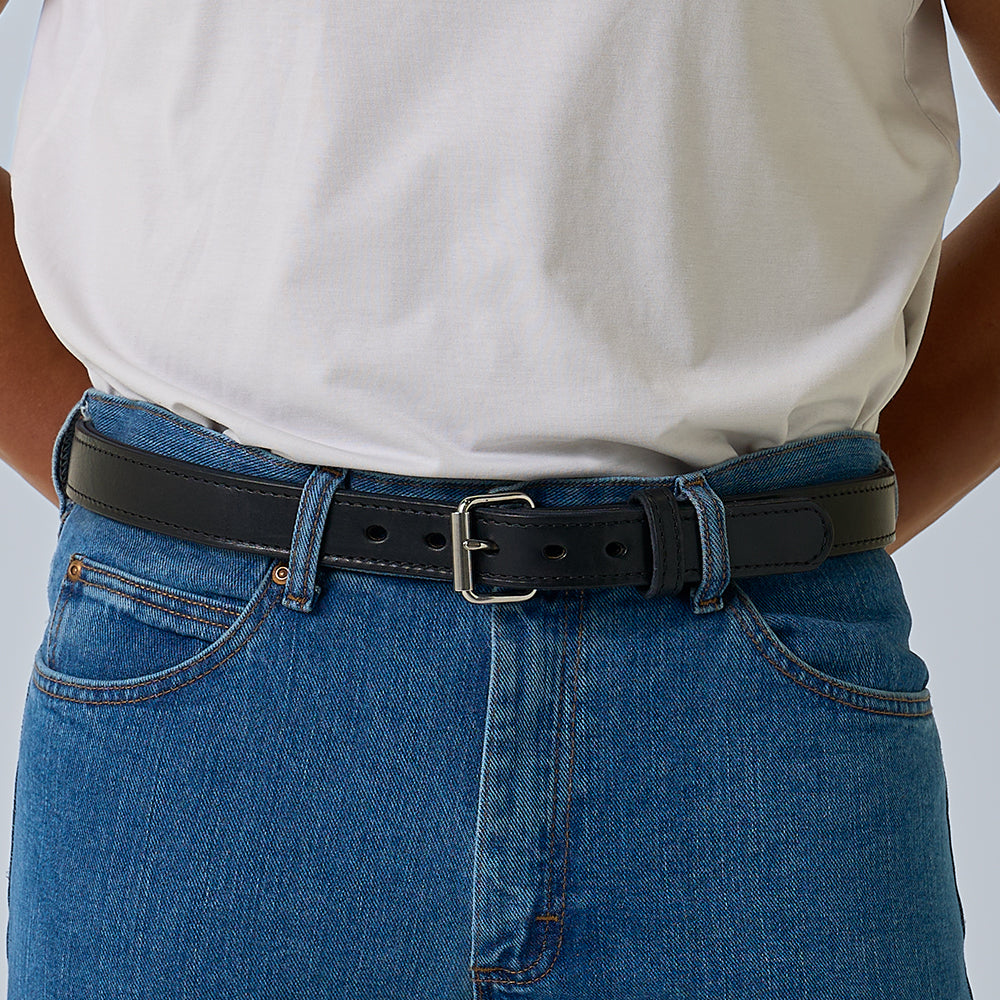 A person wearing blue jeans, a white shirt, and a Hanks Belts "The Steel Core - 1.25"" black leather gun belt fastened around the waist; image cropped to show only the torso and waist area.