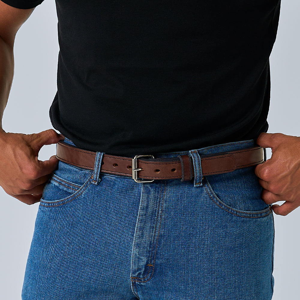 A person wearing a black t-shirt, blue jeans, and a Hanks Belts The Steel Core - 1.25" brown leather belt with a silver buckle, both hands resting near their front pockets.