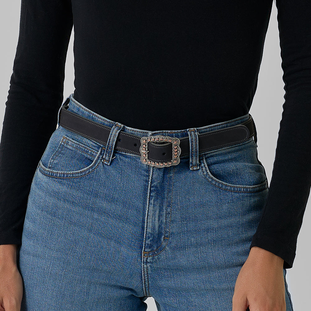 A person wears a black long-sleeve top tucked into high-waisted blue jeans, accessorized with Hanks Belts' The Lennon Suede—a stylish Western belt featuring an ornate, square silver buckle.