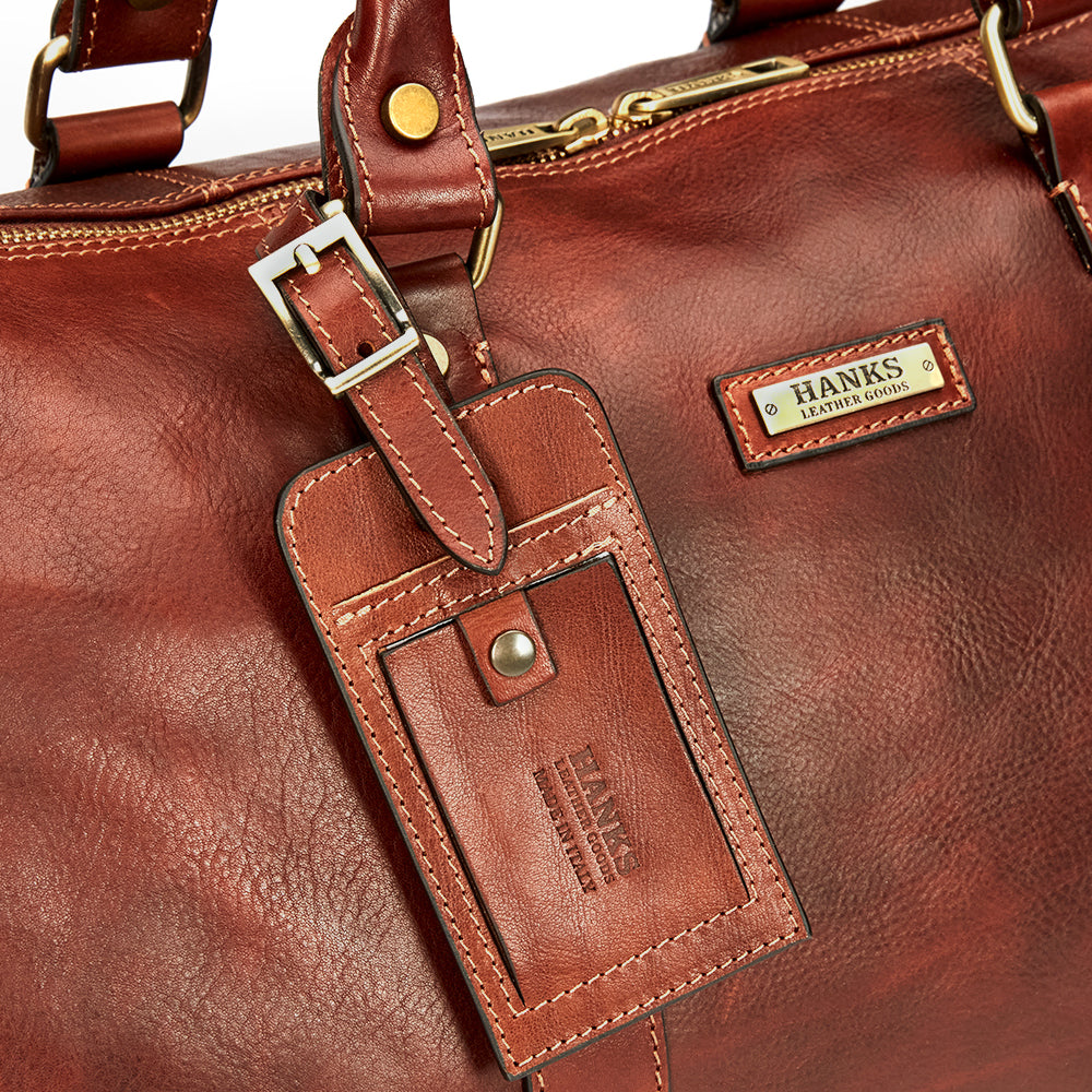 Close-up of a brown Italian-made leather bag featuring a luxury luggage tag and metal buckle. Both the tag and the bag's label are embossed with "Hanks Belts." The premium full-grain leather boasts a rich texture, visible stitching, and a golden zipper.