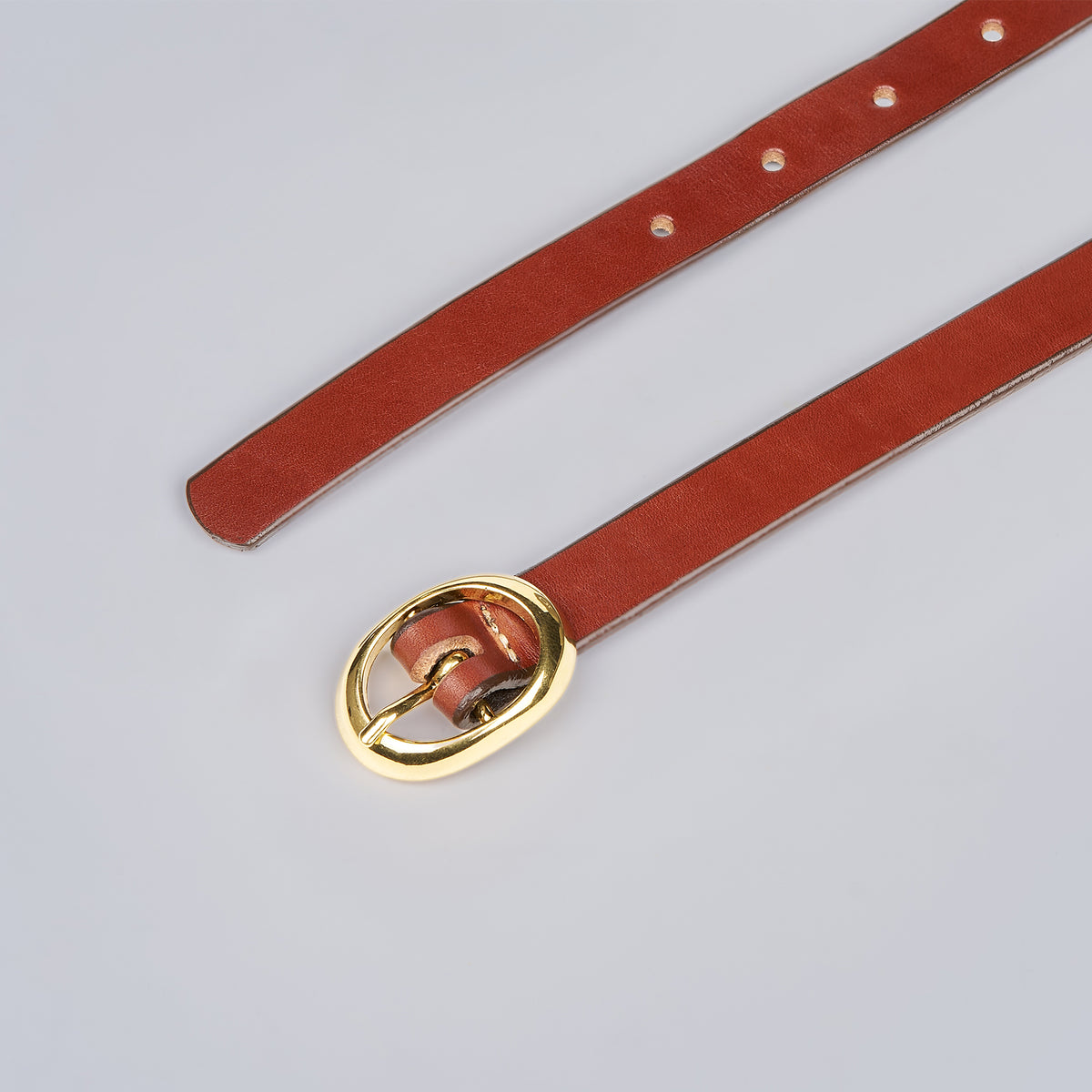 The Ivy by Hanks Belts is a brown leather belt with a shiny gold oval buckle, featuring multiple adjustment holes and a polished, smooth finish, displayed flat on a light gray surface.