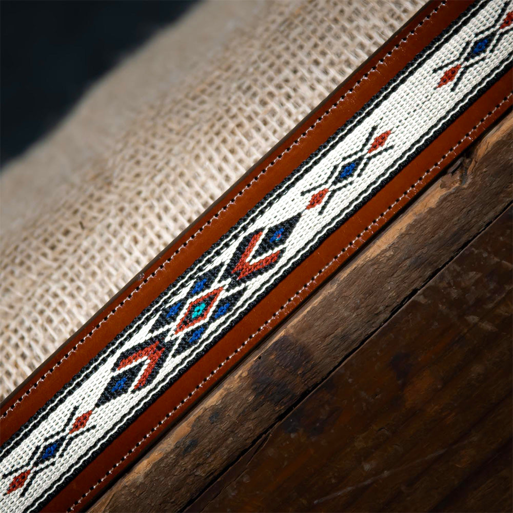 A close-up of The Seneca Tapered by Hanks Belts, made from premium English Bridle leather and featuring a colorful geometric woven pattern with diamonds and arrows, rests on burlap and wood for a western-inspired touch.