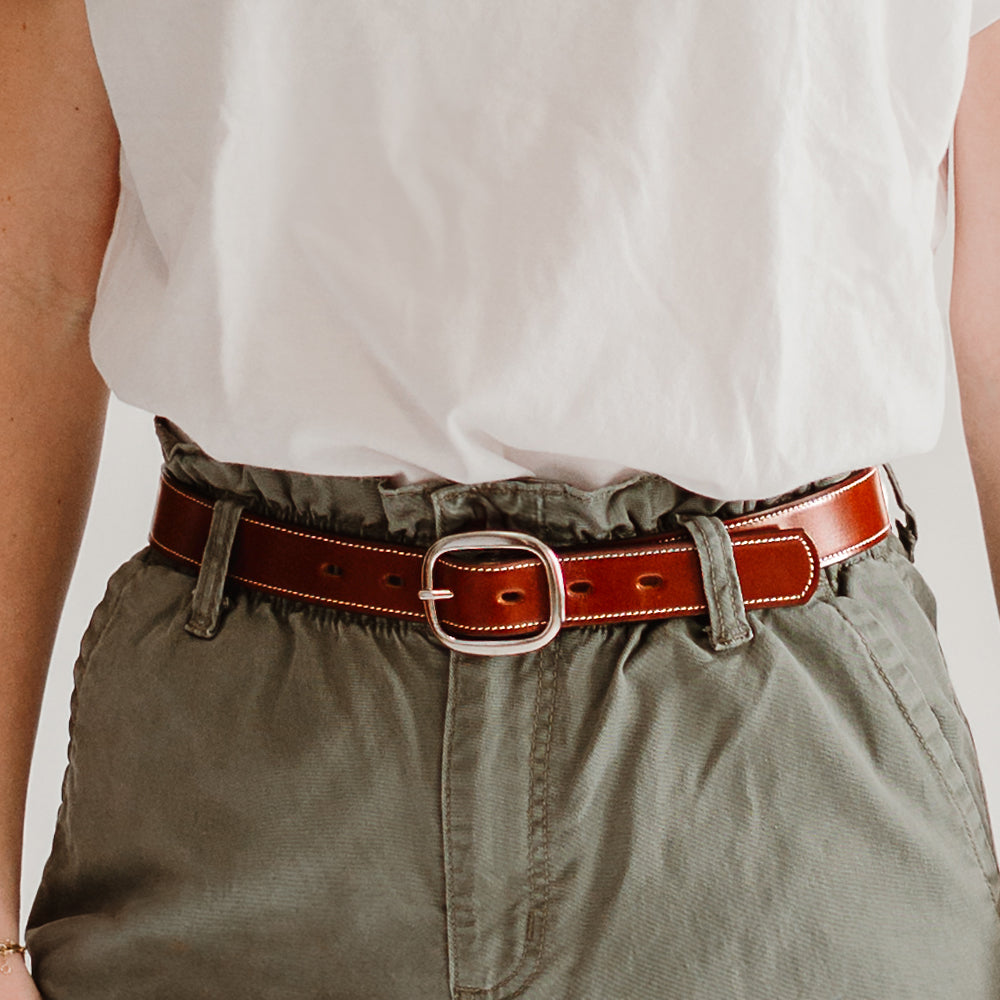 A person wears a white shirt with high-waisted olive green pants, accessorized by The Eloise—Hanks Belts’ premium vegetable-tanned leather belt with a sleek center bar buckle.