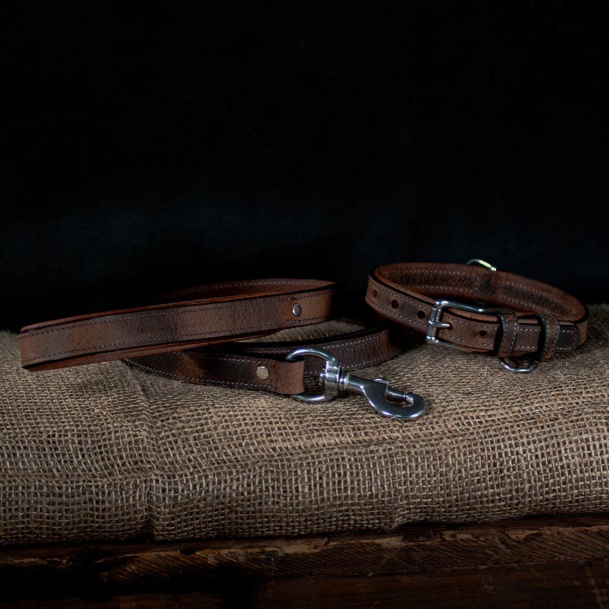 The Hanks Belts brown premium bison leather dog collar and matching Kodak Bison Leash with stainless steel hardware are displayed on textured burlap against a dark background.
