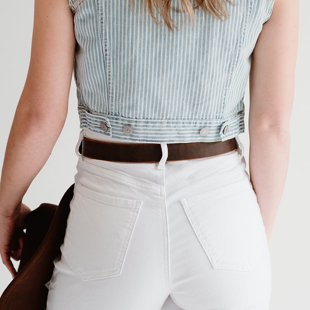 A person in a light blue striped sleeveless denim top and high-waisted white jeans, accessorized with The Lainey from Hanks Belts featuring an antique copper buckle, stands back to the camera holding a brown hat.