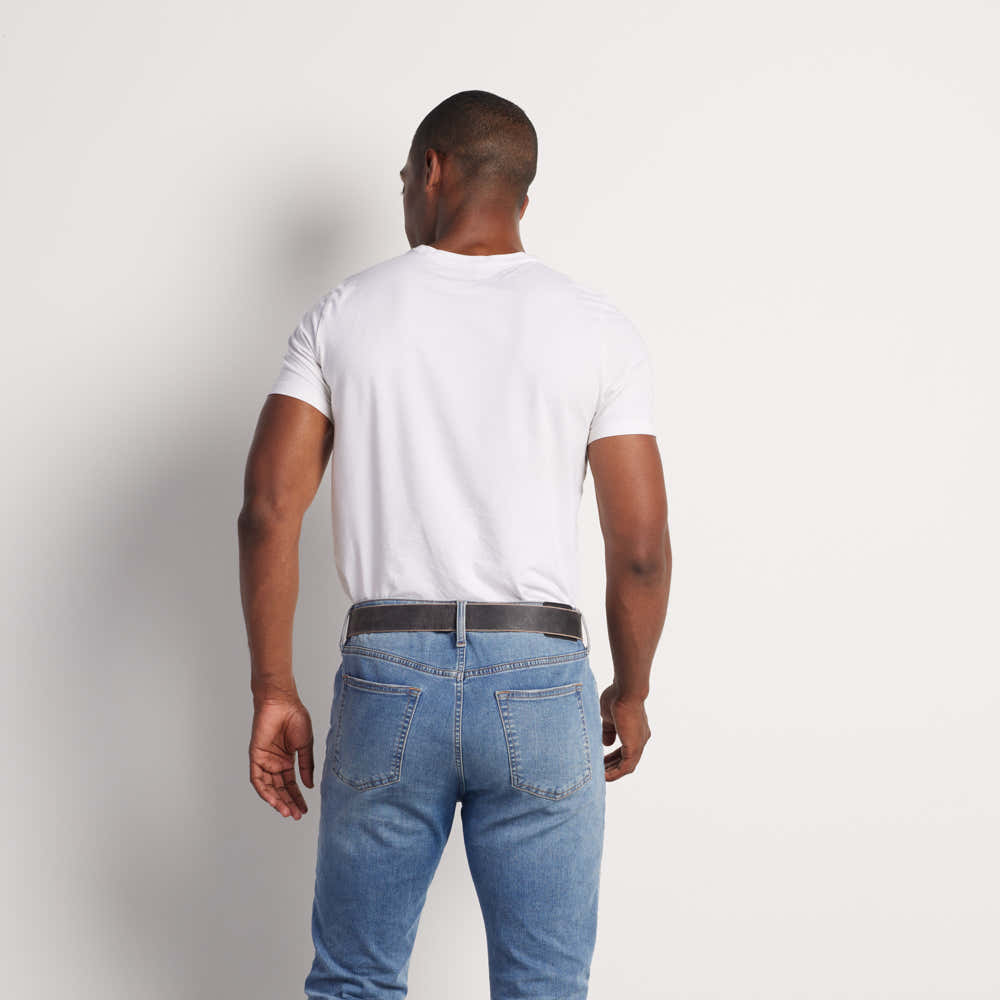A man stands with his back to the camera, wearing a white t-shirt, blue jeans, and The Glacier Grey Retro Style belt by Hanks Belts—crafted from USA-made water buffalo leather—against a plain light background.