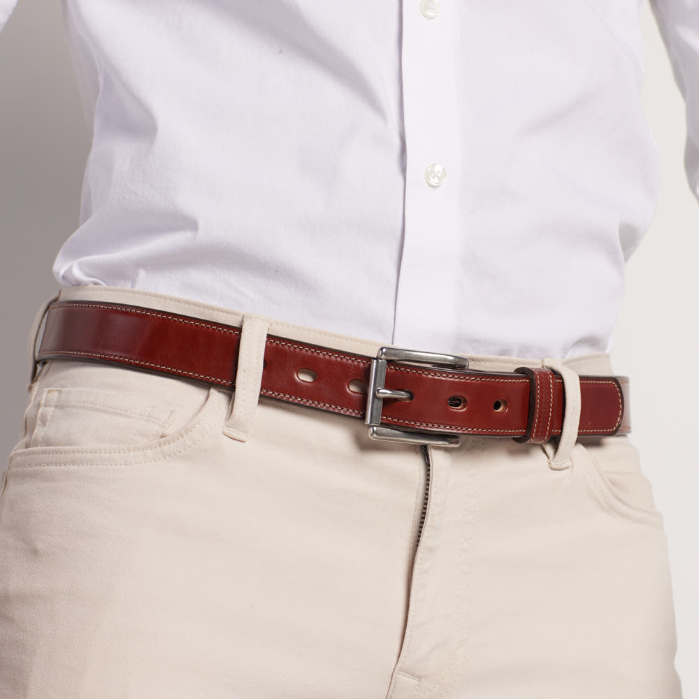A person wears a white button-up shirt tucked into light beige pants, paired with Hanks Belts’ The Highland Lined—a shiny English bridle leather belt finished with a silver buckle.