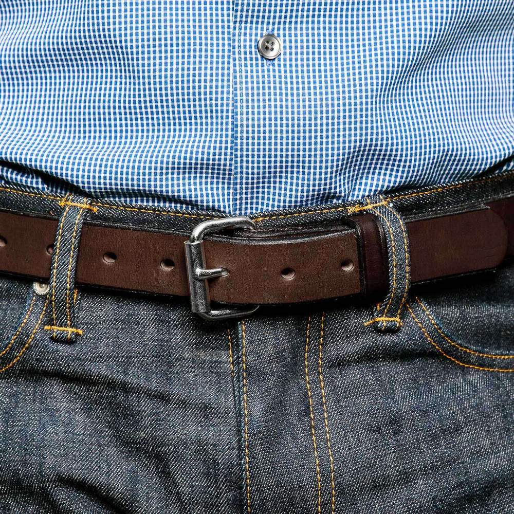 Wearing a blue checkered shirt tucked into blue jeans with yellow stitching, the person secures their look with The Deputy by Hanks Belts, featuring a durable leather strap and metal buckle.