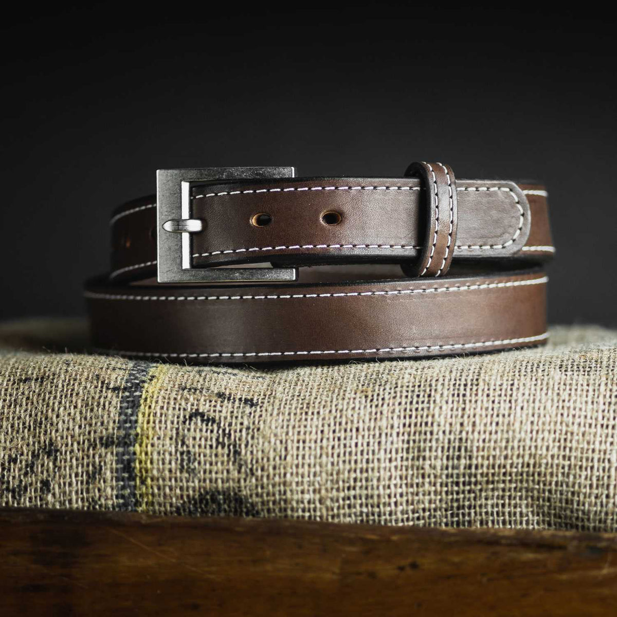 The Bonnie by Hanks Belts, made from full-grain vegetable tanned leather with white stitching and a silver buckle, is coiled neatly on a textured fabric surface against a dark background.
