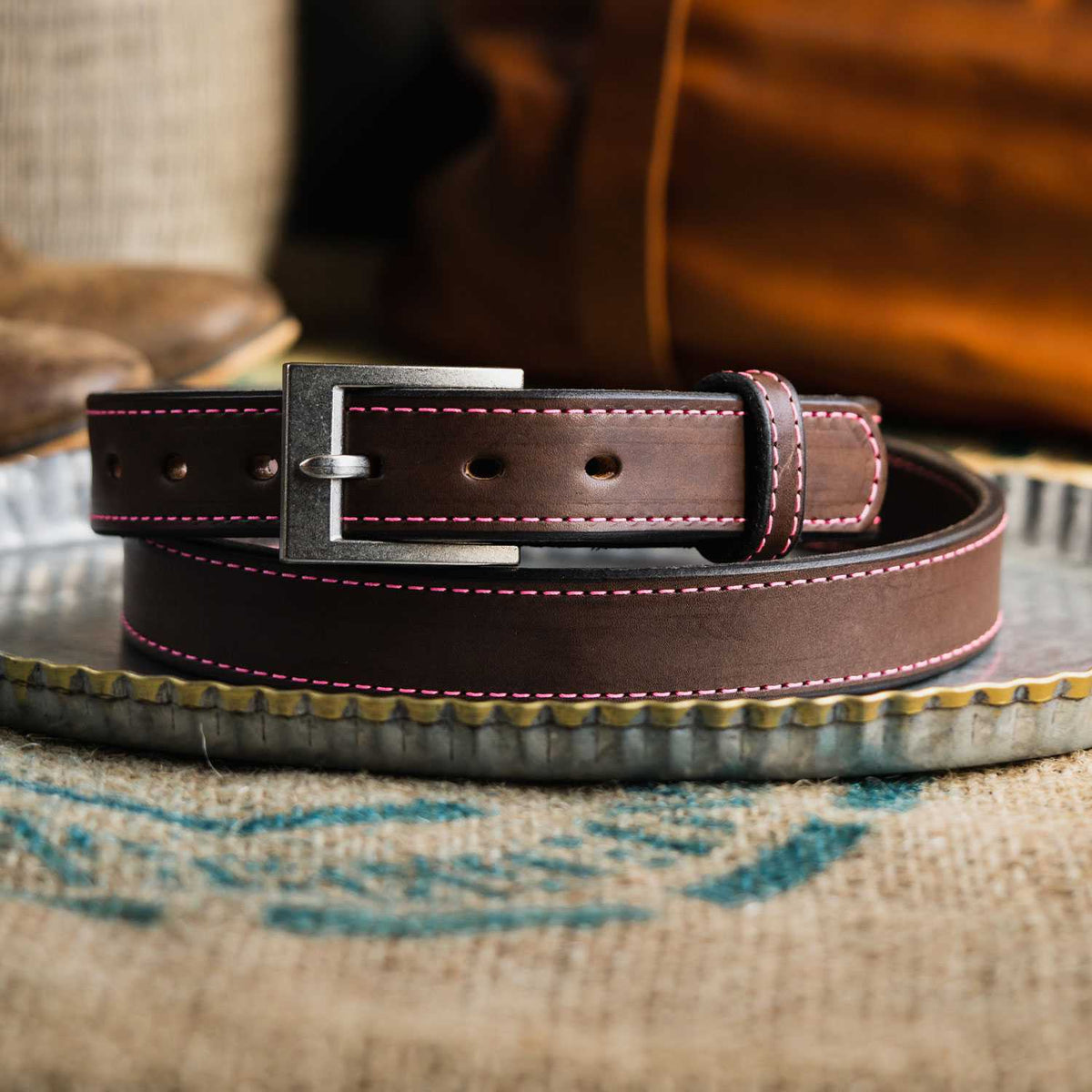 The Bonnie by Hanks Belts, made from full-grain vegetable tanned leather with red stitching and a silver buckle, is coiled on a corrugated metal tray with blurred fabric and leather items in the background.