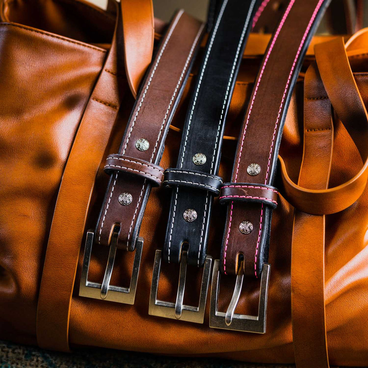 Three "The Bonnie" belts by Hanks Belts—two brown, one black—crafted from full-grain vegetable tanned leather with metal buckles and white or red stitching, are displayed side by side on a large brown leather bag.