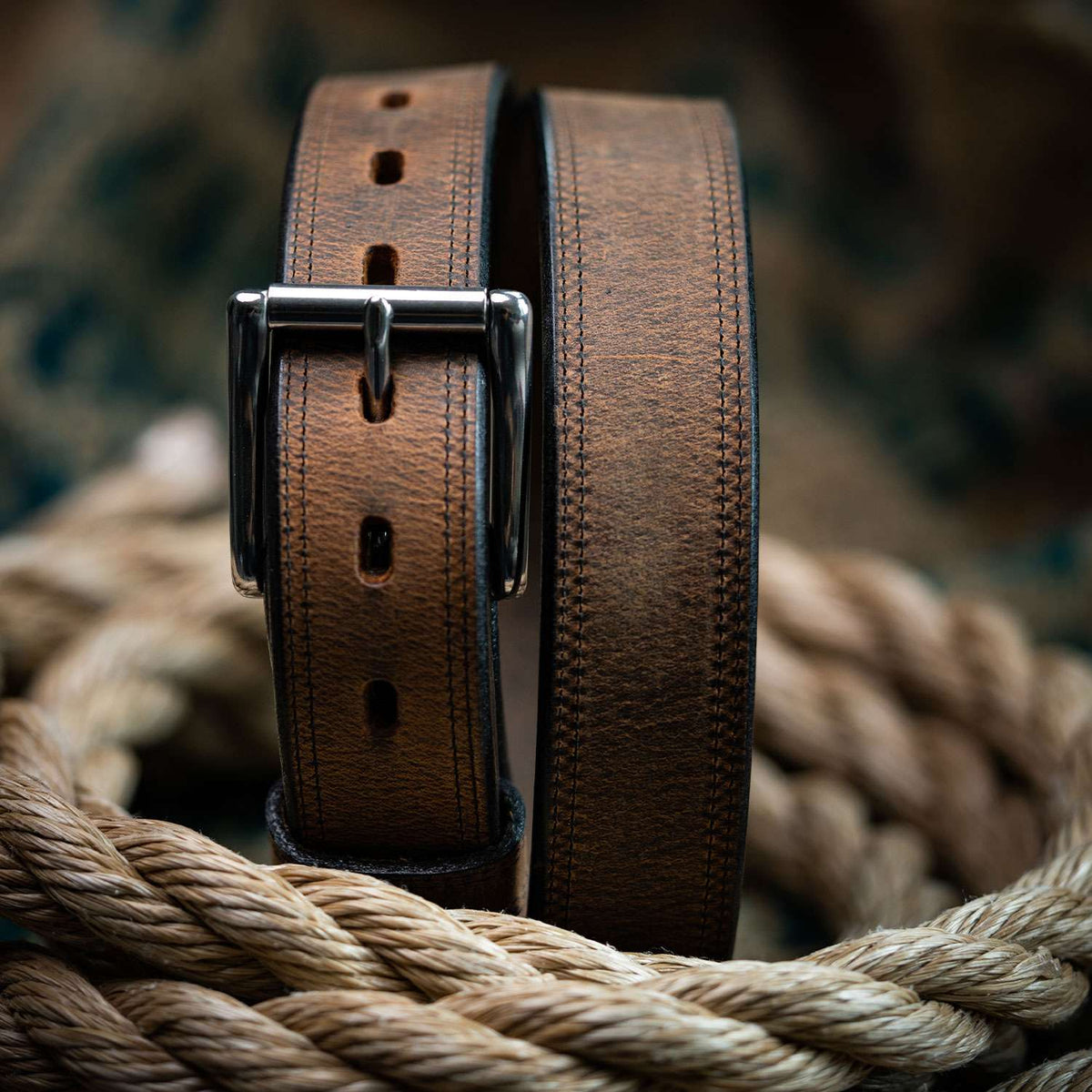 A close-up of The Montana Bison Double Layer by Hanks Belts, a rugged brown bison leather belt with a stainless steel buckle, coiled on thick beige rope against a blurred background.