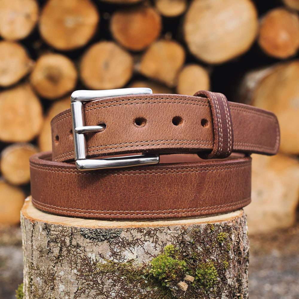 The Montana Bison Double Layer by Hanks Belts, crafted from rugged bison leather with a stainless steel buckle, rests coiled on a mossy stump as stacked logs blur in the background.