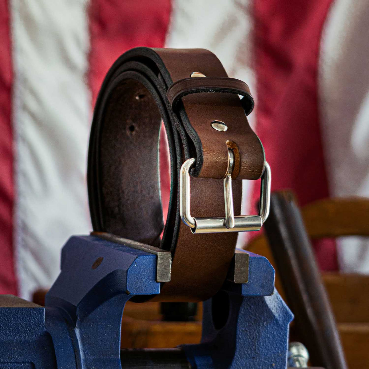 The Enforcer full-grain leather belt by Hanks Belts, featuring a silver buckle, is displayed on a blue metal vise with a blurred red and white striped background.