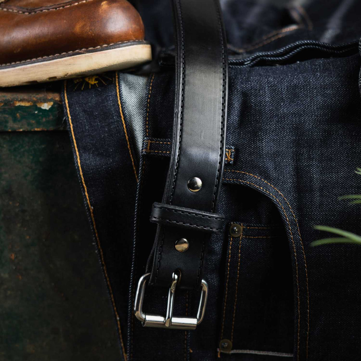 A close-up of a pair of dark blue denim jeans with contrast stitching, laid flat with a black leather Hanks Belts Steel Core Belt - 1.5" featuring a silver buckle draped across them. The edge of a brown leather boot is visible at the top left corner of the image.