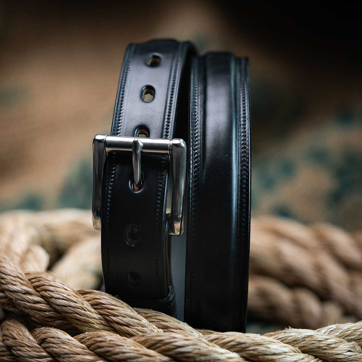 A close-up of The Esquire Raised belt by Hanks Belts, crafted from English Bridle leather with a silver buckle, coiled atop thick braided rope against a blurred background.
