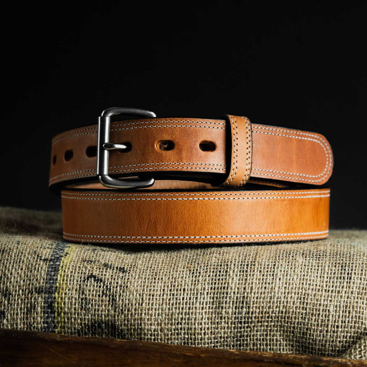 The Old World Harness Leather Belt - 1.5" by Hanks Belts, crafted from USA-tanned Hermann Oak full-grain leather with white stitching and a metal buckle, is coiled on burlap fabric against a black background.