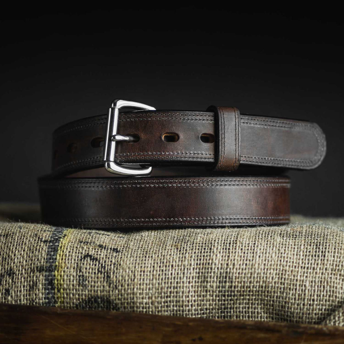 The Old World Harness by Hanks Belts, a dark brown full-grain leather belt with a silver buckle, is coiled atop a textured burlap surface against a dark background.