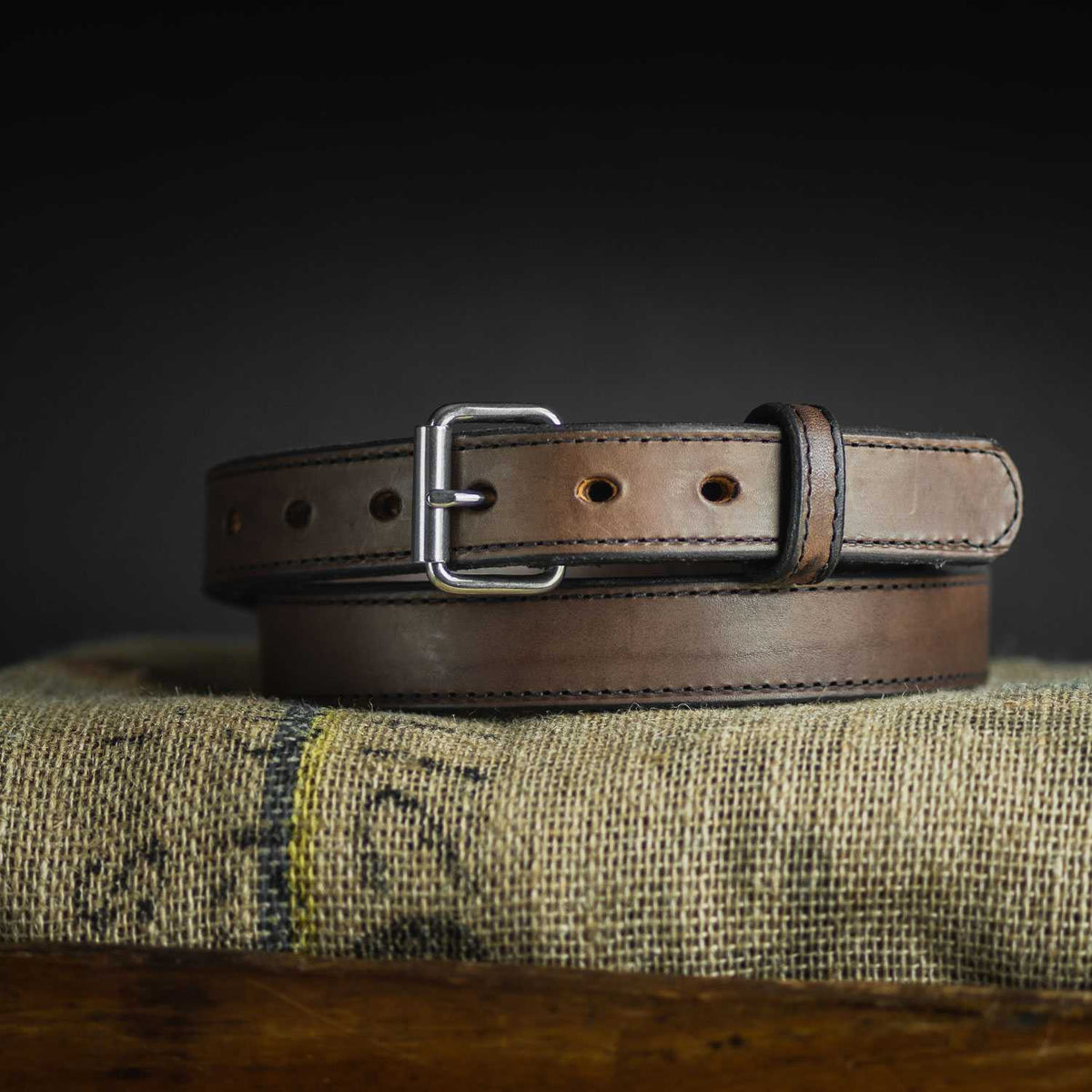 A Hanks Belts "The Steel Core" brown leather gun belt with black stitching and a silver buckle is coiled on textured burlap against a dark background.