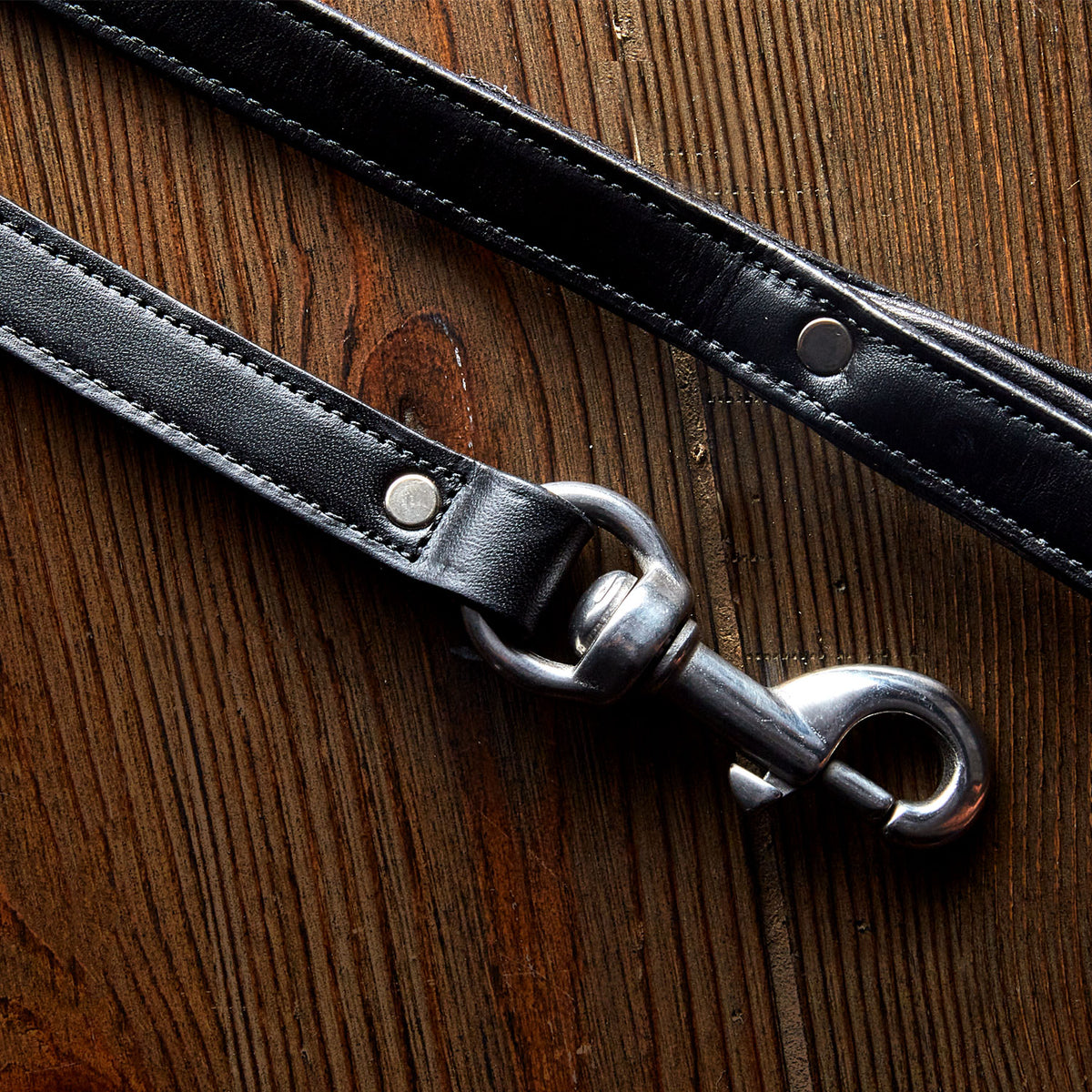 A close-up of the Rover Standard Leash by Hanks Belts, crafted from black English Bridle leather with a silver clasp and padded handle, displayed on a wooden surface.