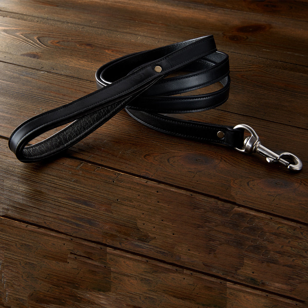 The Hanks Belts Rover Standard Leash, featuring black leather, a padded handle, and sturdy metal clasp, is coiled on a dark wooden surface.