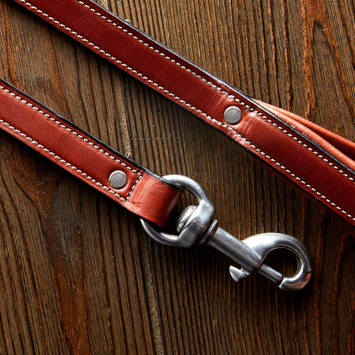 A close-up of the Hanks Belts Rover Standard Leash in brown English Bridle leather with white stitching, padded handle, and metal hardware, resting on a wooden surface.
