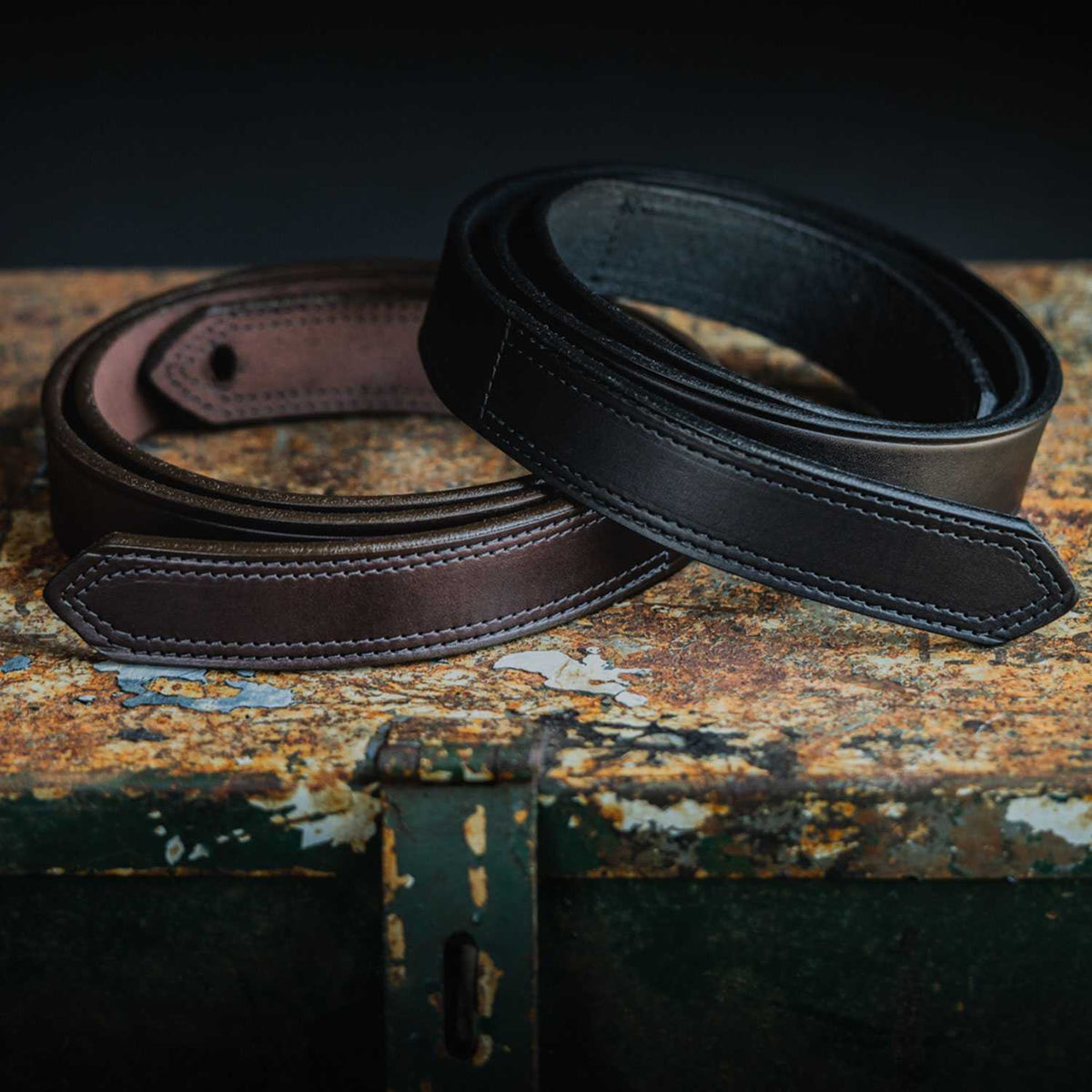 Two Hanks Belts "The Scratchless Mechanics" belts, one brown and one black, made from genuine full grain leather, are coiled and displayed on a rustic metal surface against a dark background.